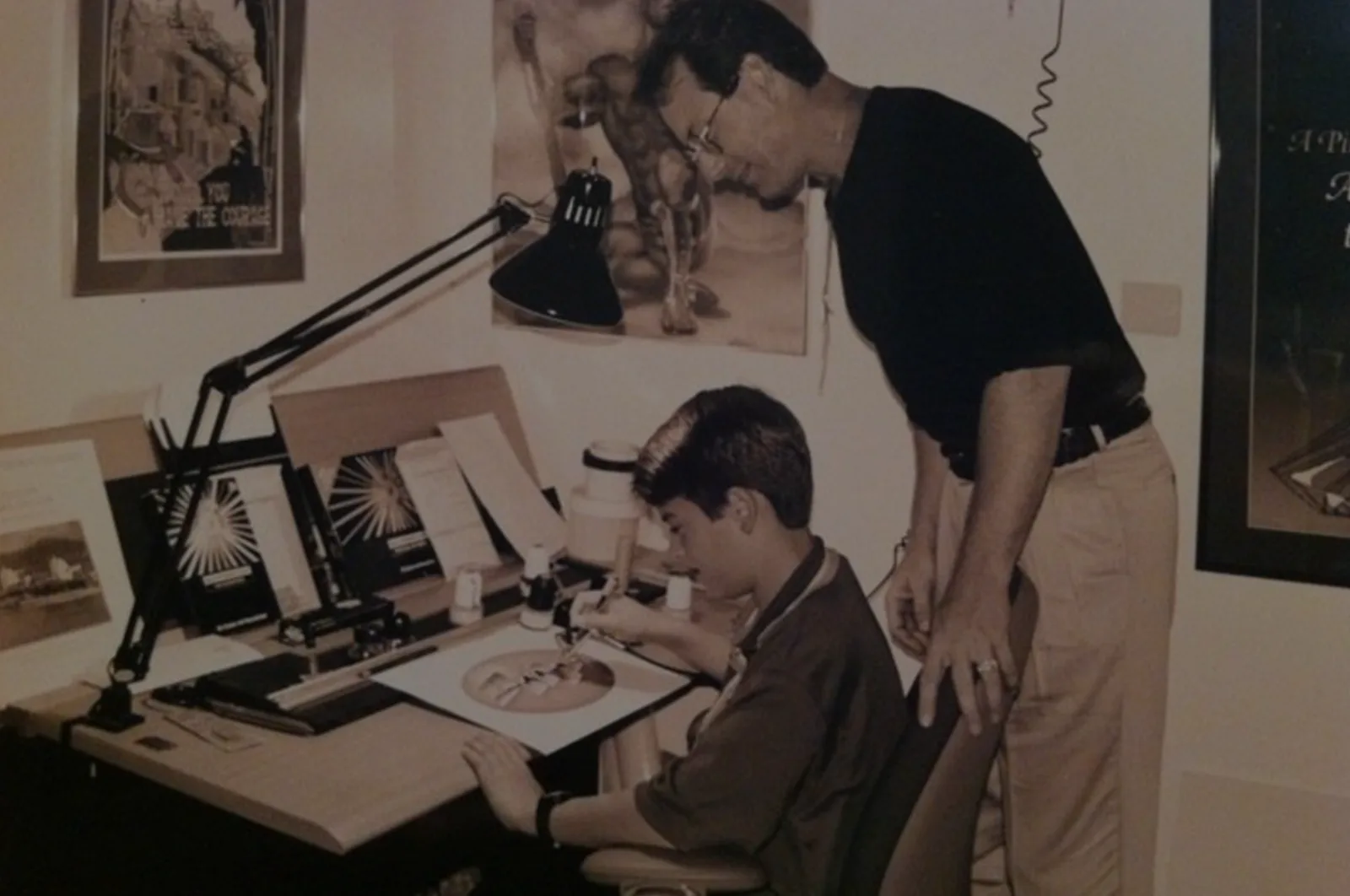 A young Michael working at a drafting table under an architect's lamp while his dad leans in over his shoulder to watch.